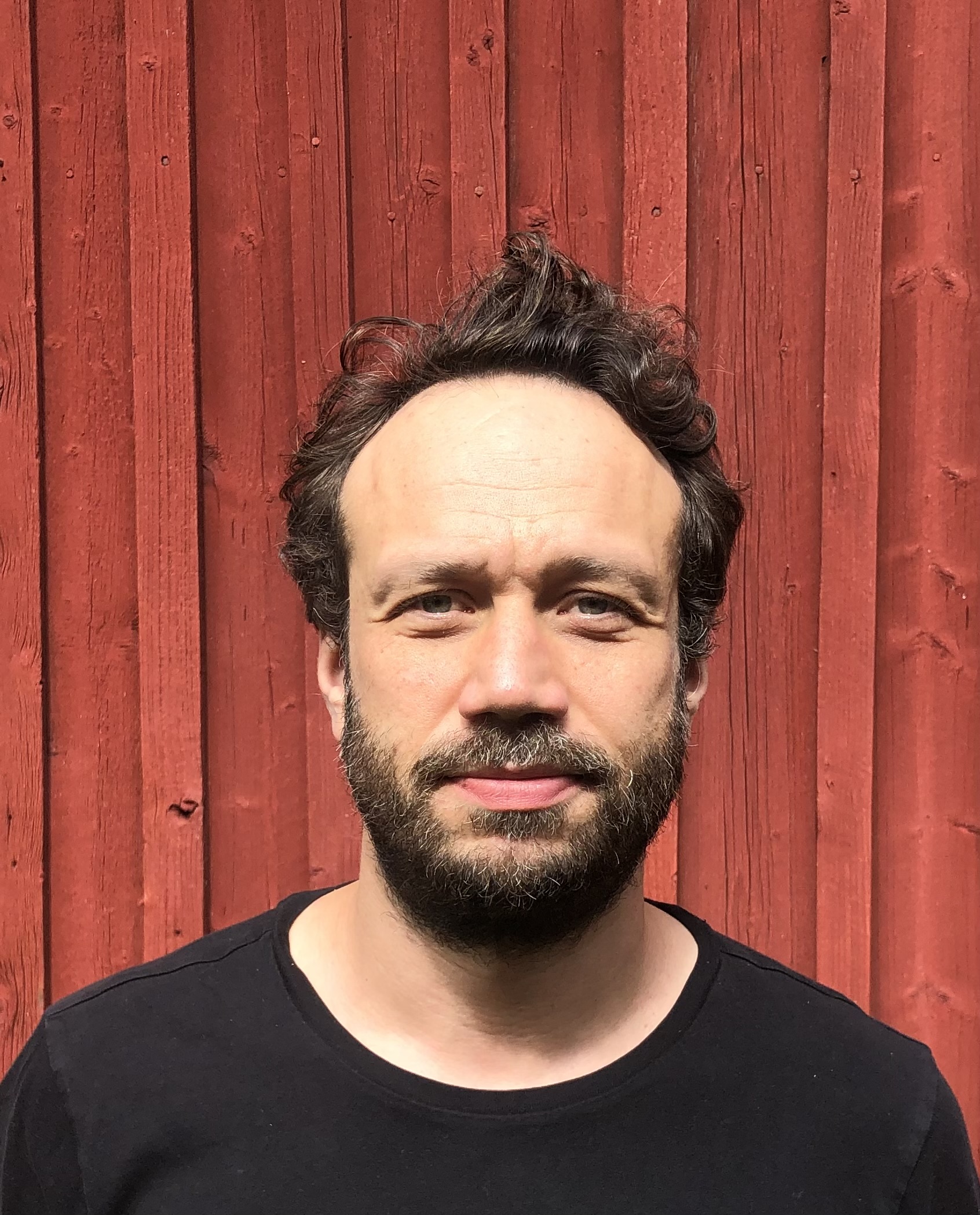 En bild på en man med brunt hår och skägg som står mot en röd vägg.